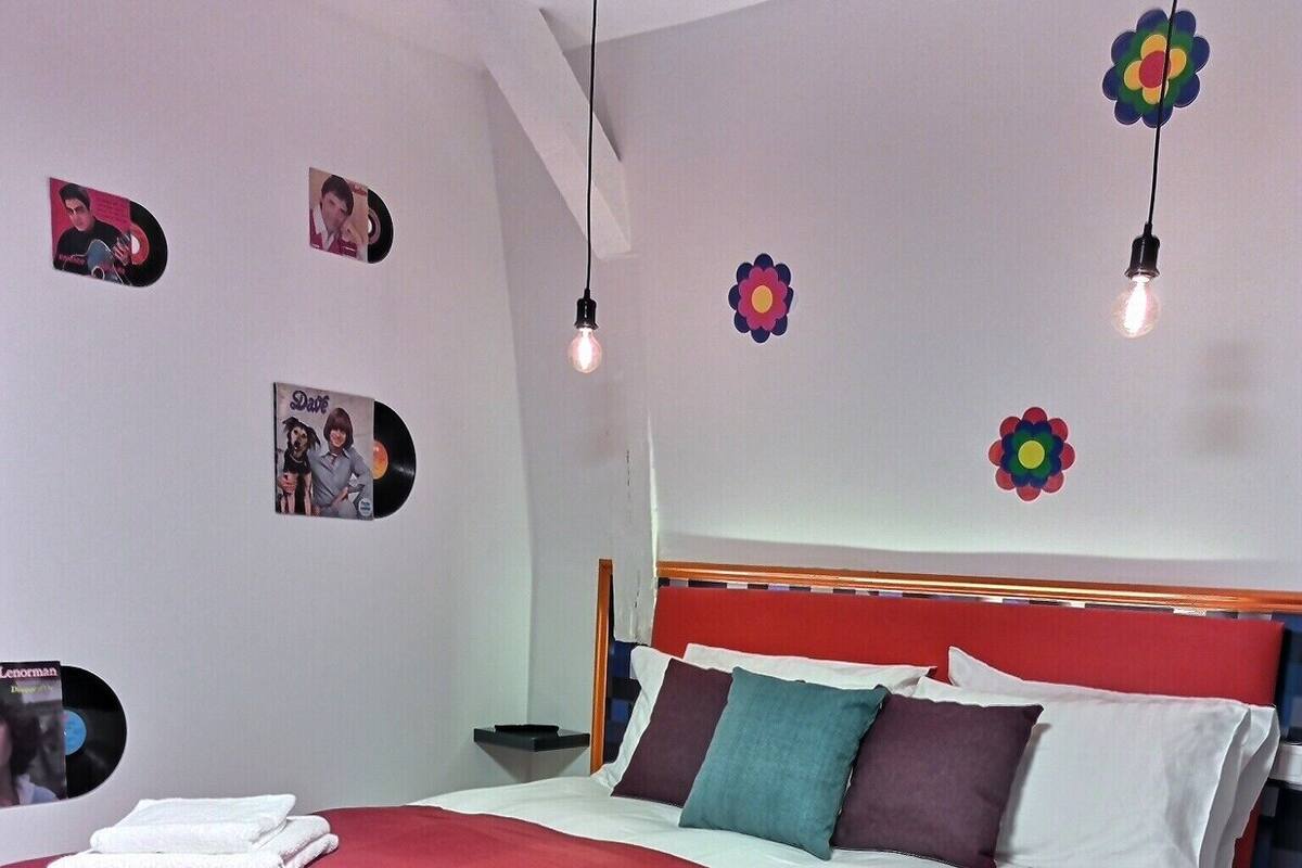 Una cama bien hecha con una colcha roja, almohadas blancas y un cabecero de madera. Hay dos adornos florales en la pared y algunos discos de vinilo también colgados allí.