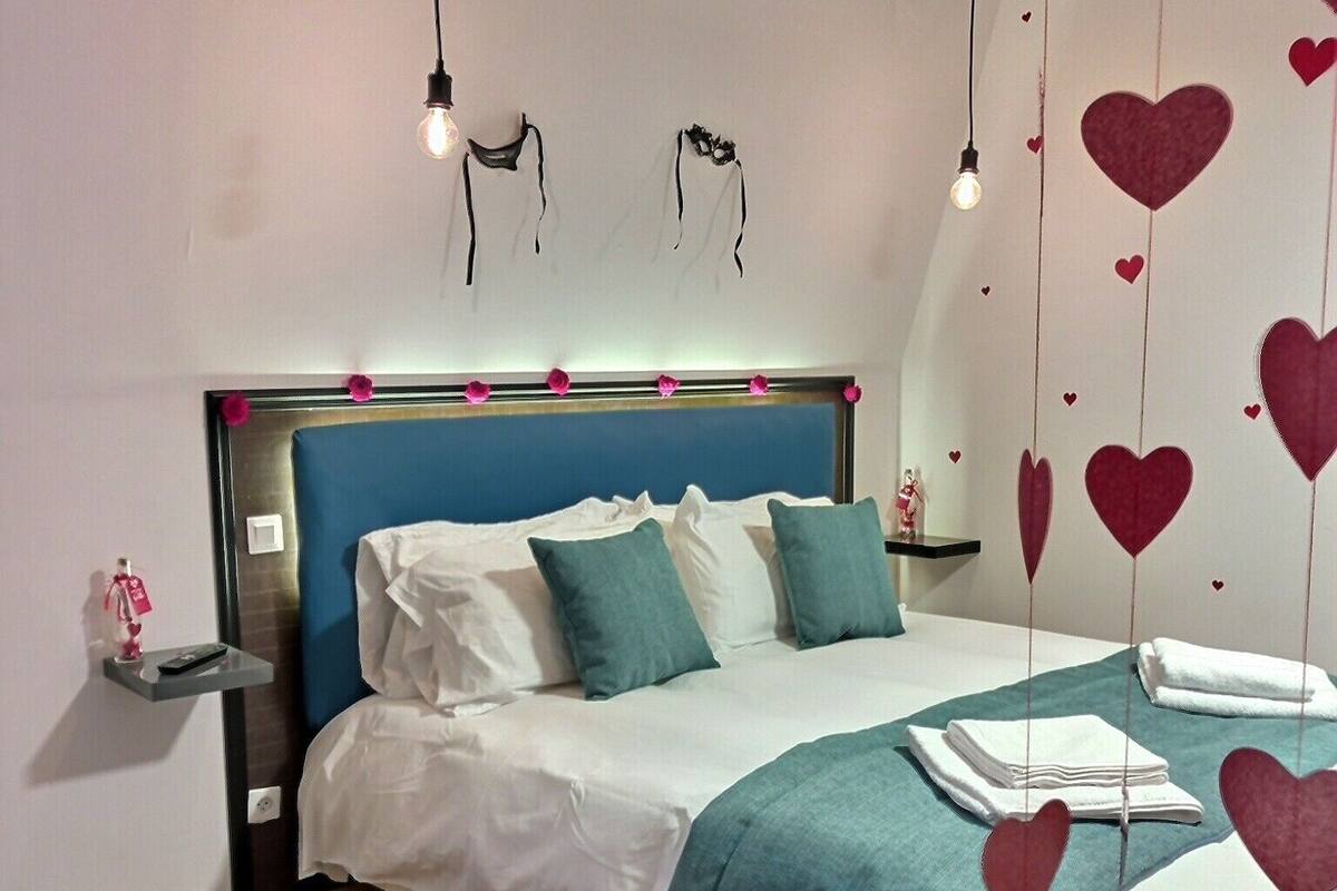 Un dormitorio con cabecera azul, ropa de cama blanca, almohadones color turquesa, alfombra roja y adornos con forma de corazón en la pared.
