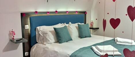 A bedroom with a blue headboard, white bedding, teal throw pillows, a red rug, and heart-shaped decorations on the wall.