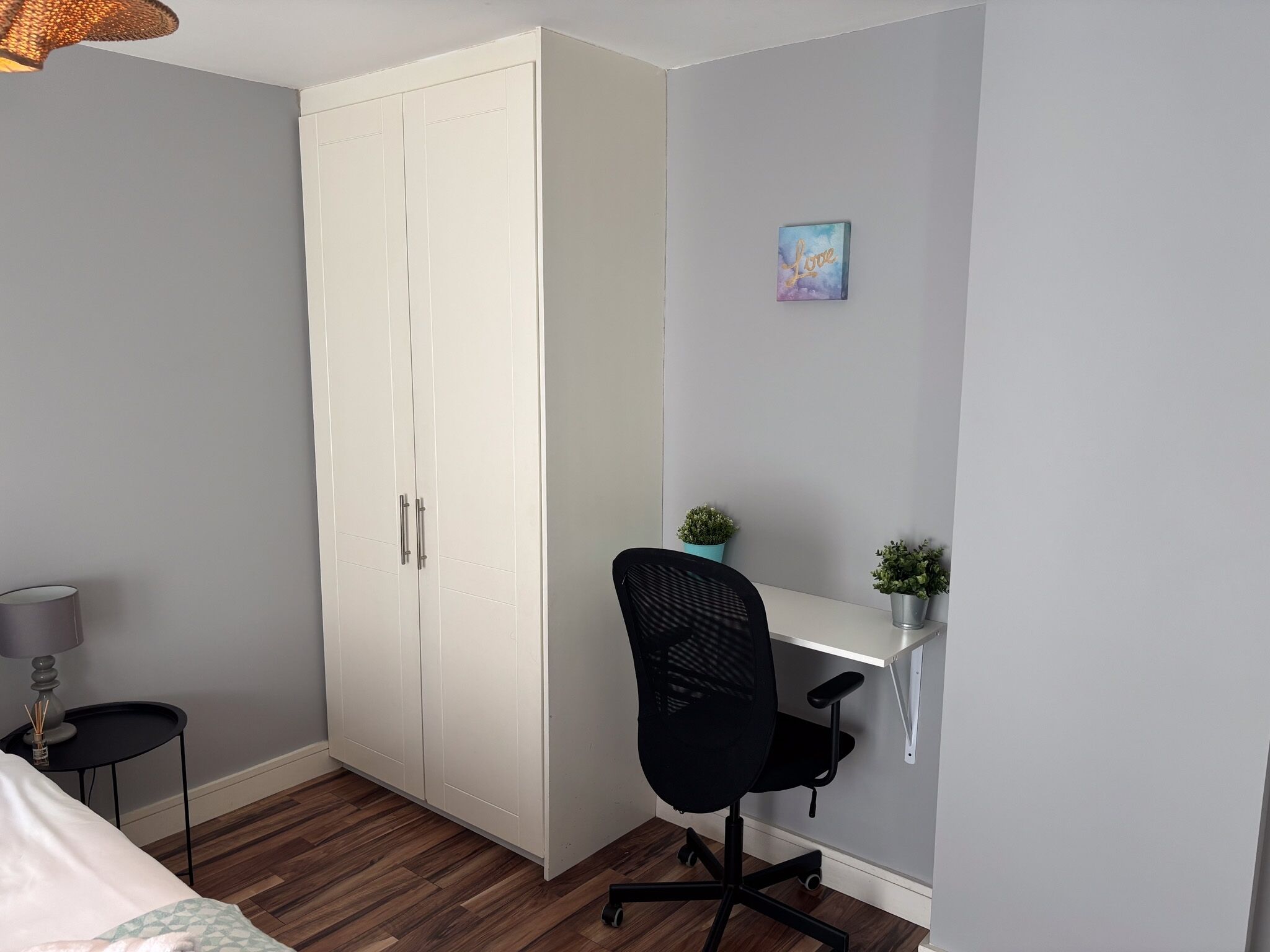 Ett sovrum med en säng, en garderob, ett skrivbord med en stol, en lampa och en liten växt.