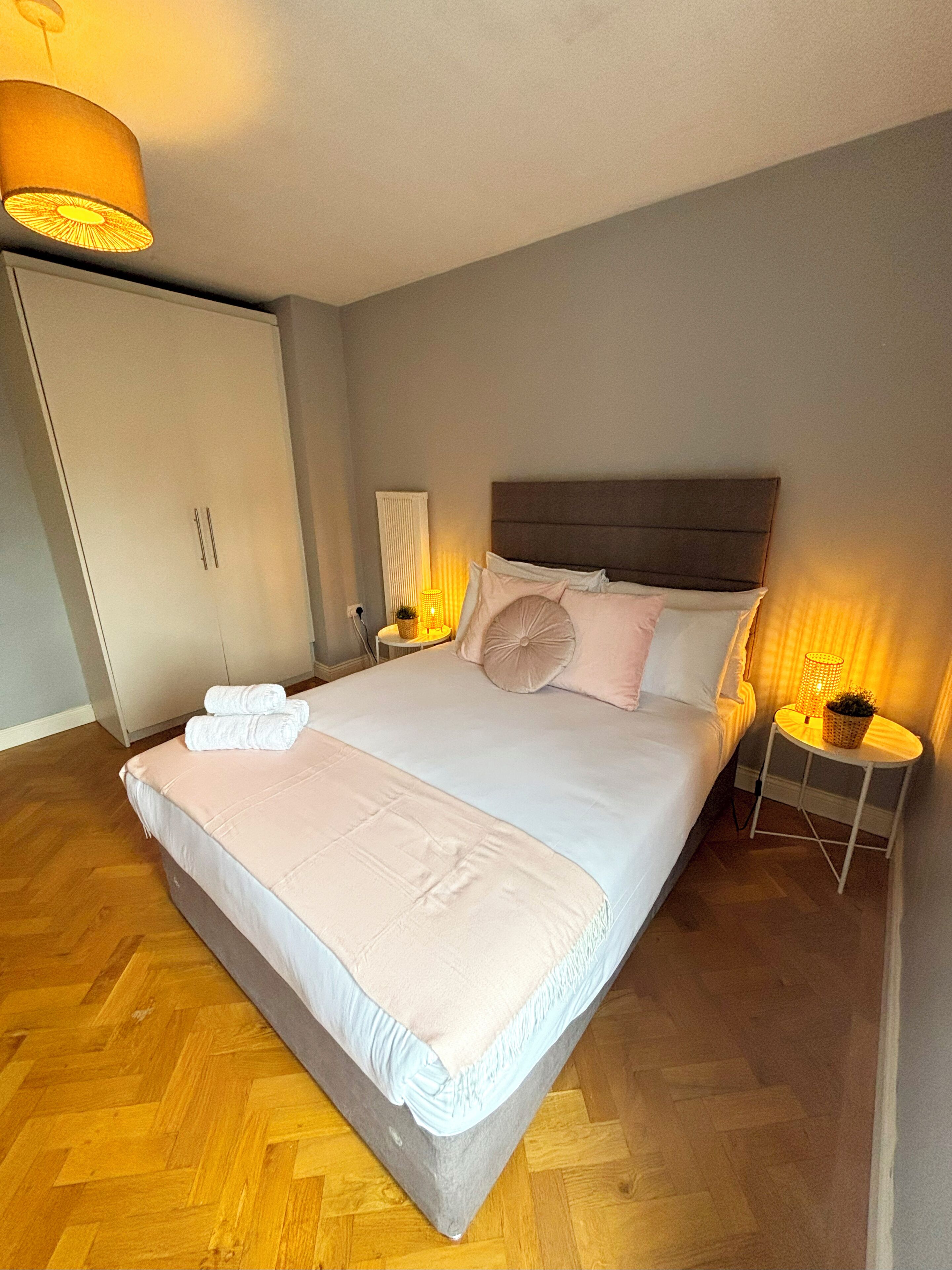 Una cama bien hecha con almohadas y una colcha, una mesita de noche con lámpara, un armario y un radiador.