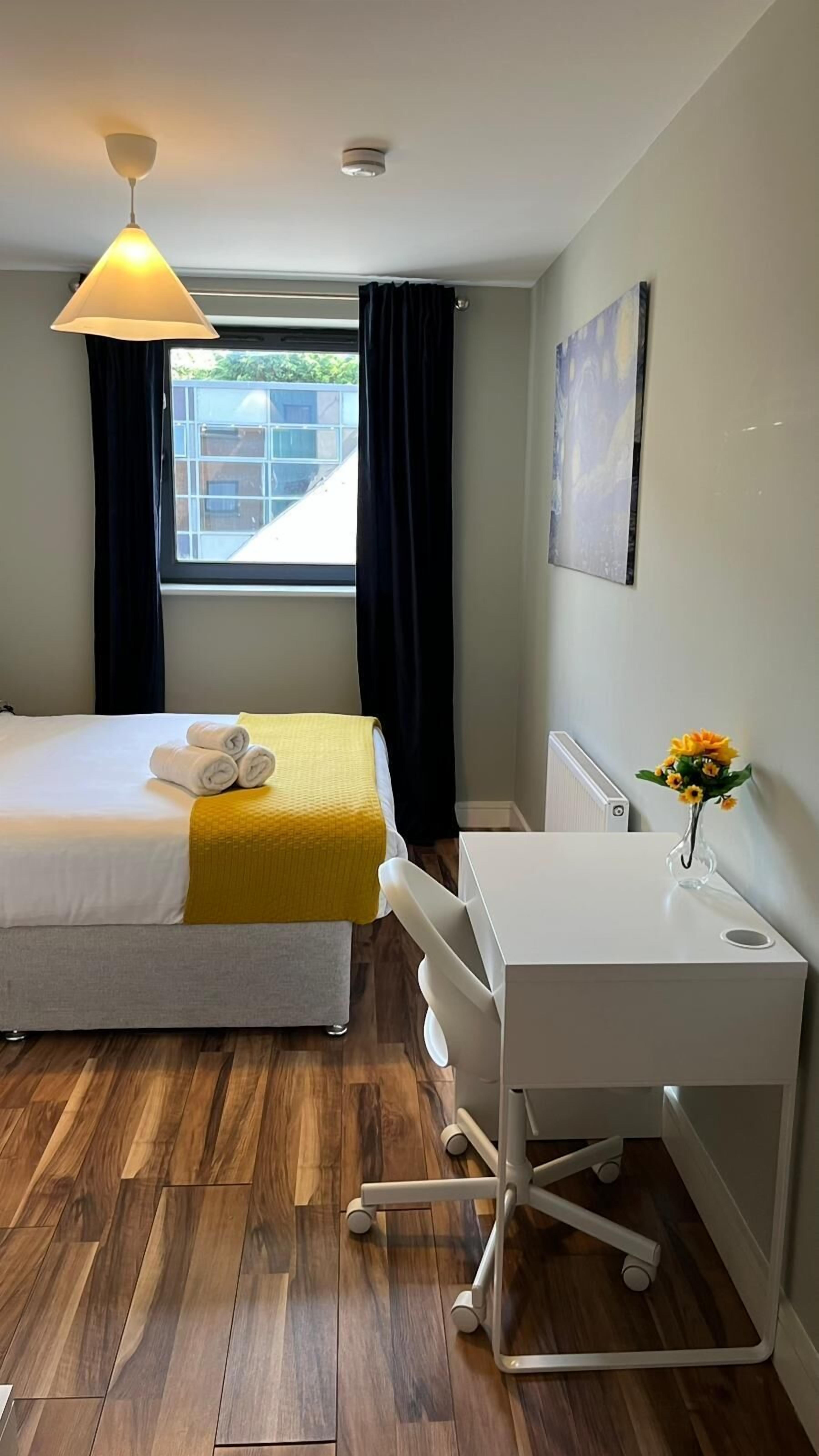 A hotel room with a bed, a desk, a chair, a window with curtains, a ceiling light, and a radiator.