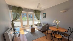 Un salón moderno con sofá, mesa de centro, mesa de comedor y sillas. Se aprecia un radiador, una ventana con cortinas e imagen colgada en la pared.