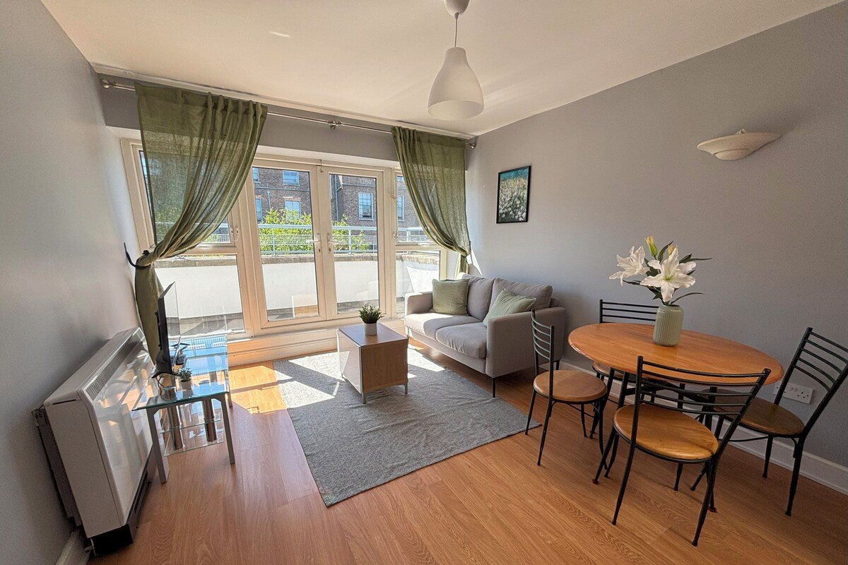 A modern living room with a sofa, coffee table, dining table, and chairs. There is a radiator, a window with curtains, and a picture on the wall.