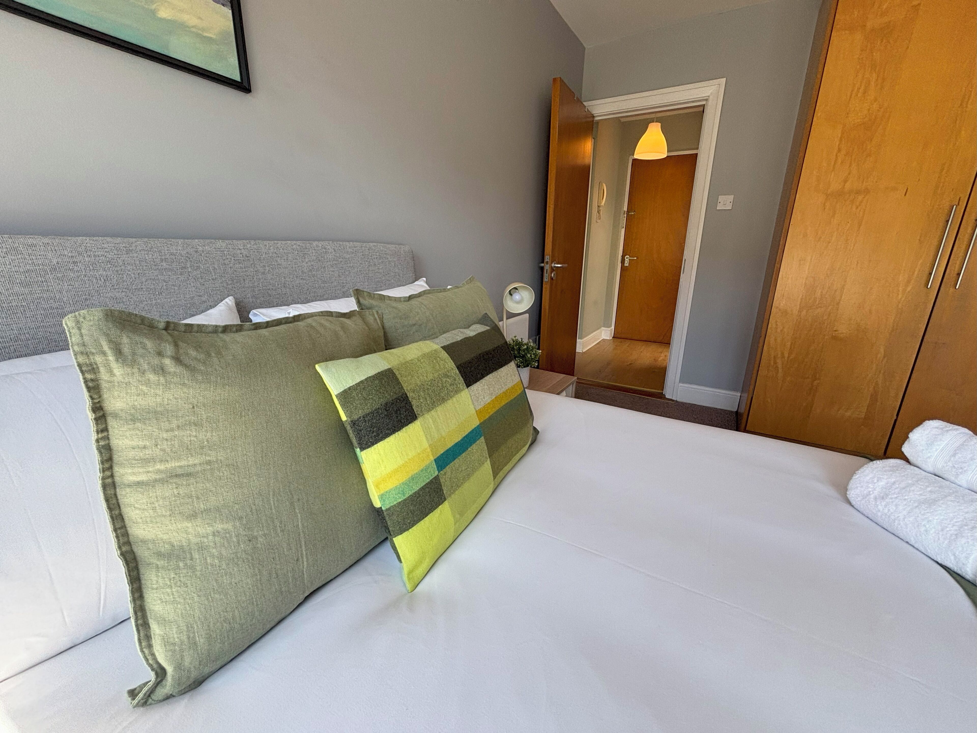 Een hotelkamer met een bed, kussens, een nachtkastje, een lamp en een houten garderobe.