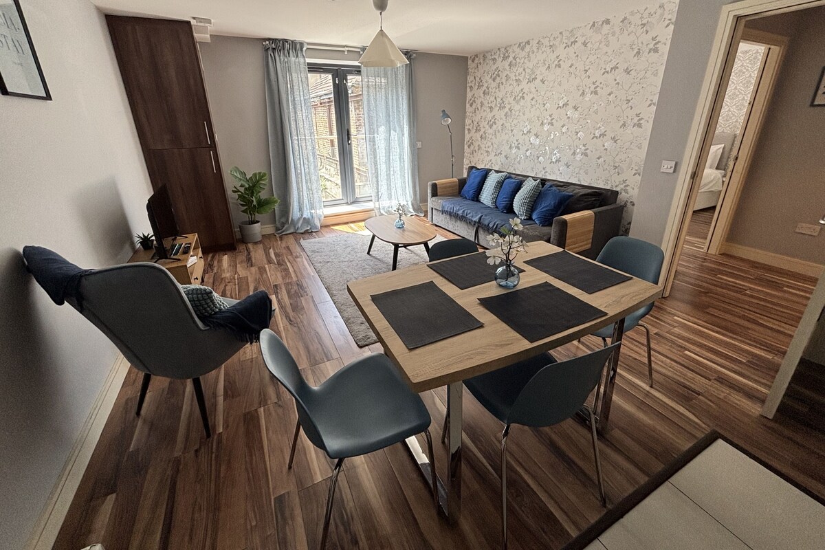 A modern living room with a dining area, wooden flooring, a sofa with blue cushions, a ceiling fan, and a large window with curtains.
