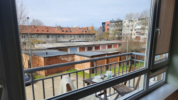 A balcony with a view of a residential area, including buildings with red roofs and brick structures. - Home-spirit apartment with a panoramic window in the heart of Klaipėda! (Klaipeda)