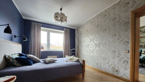 A bedroom with a bed, a nightstand, a reading lamp, a window with curtains, and a patterned wallpaper. - Home-spirit apartment with a panoramic window in the heart of Klaipėda! (Klaipeda)
