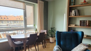 A modern living room with a dining area, a bookshelf, and a large window offering a view of buildings. - Home-spirit apartment with a panoramic window in the heart of Klaipėda! (Klaipeda)