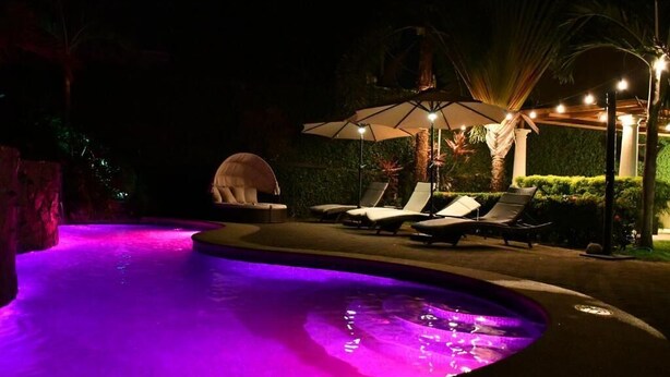Ein Schwimmbad bei Nacht mit violetter Beleuchtung, Liegestühlen und Sonnenschirmen.