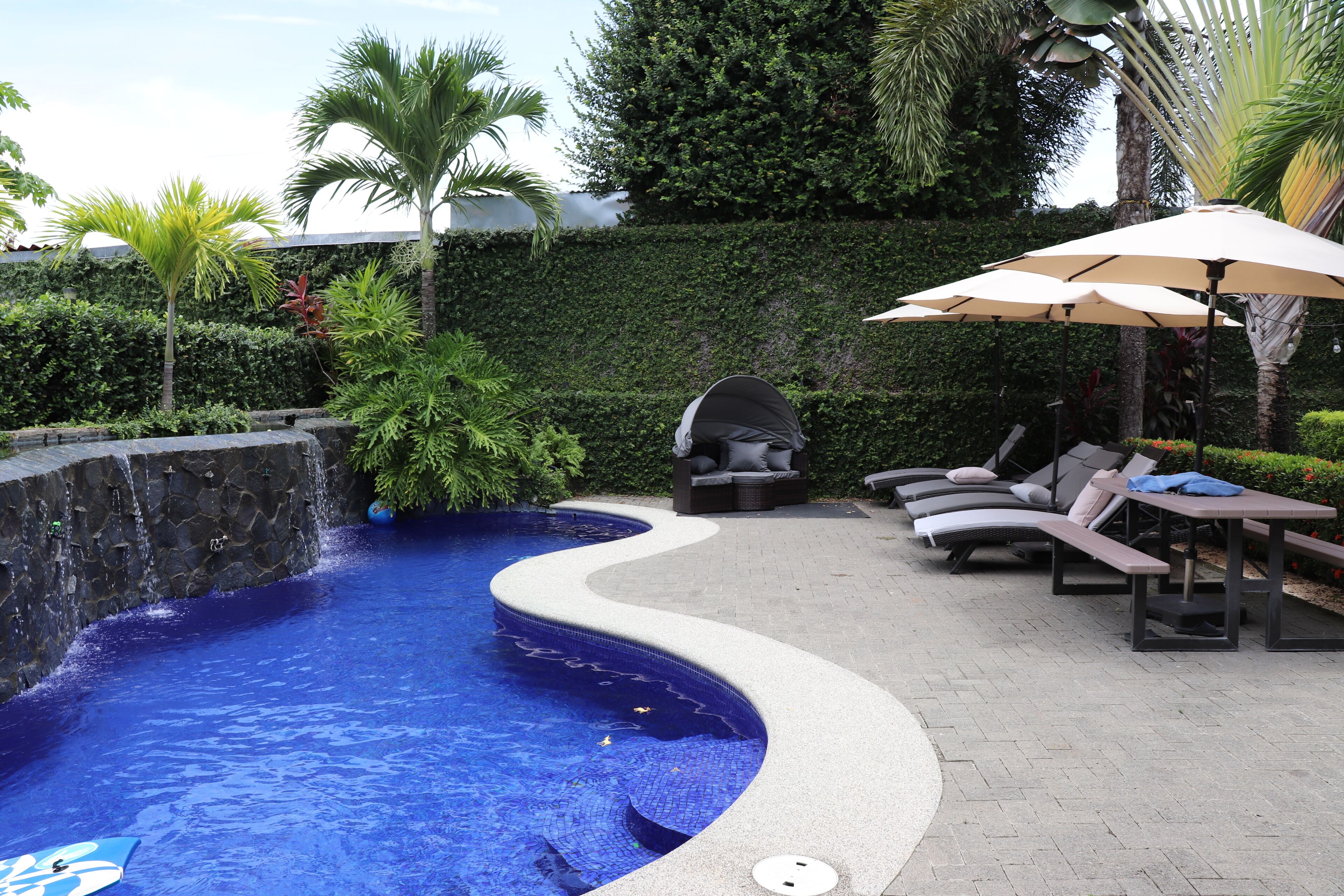 Une piscine à bord incurvé, entourée de chaises longues et de parasols, et un mur de pierre agrémenté d’un élément décoratif aquatique.