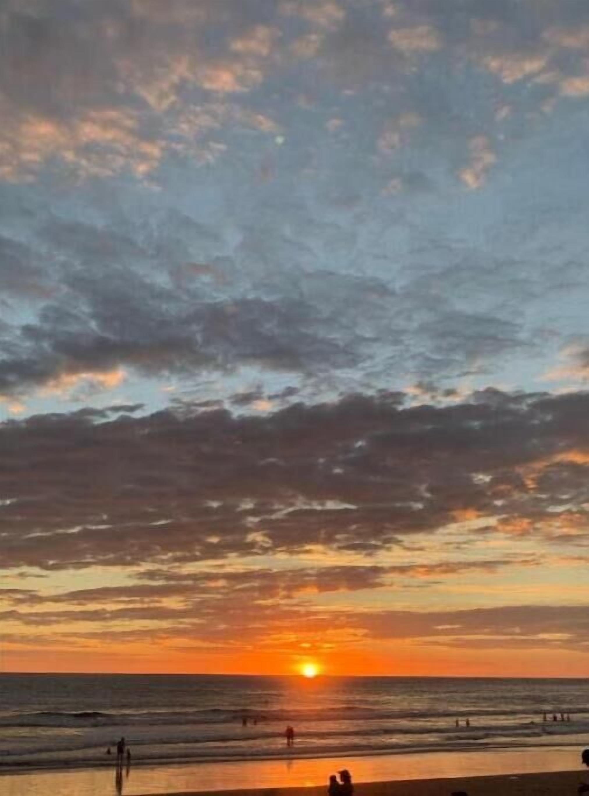 A beach scene with the sun setting over the horizon, people walking along the shore, and a clear sky.
