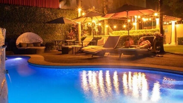 A swimming pool with lounge chairs and umbrellas, surrounded by a landscaped garden and a building with a red facade. - 12B Jaco Sol Quadplex - Luxury apartment near the beach and commerce (Jacó)