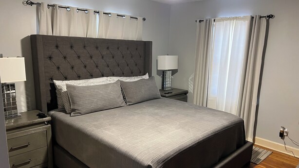 Un dormitorio con cabecera capitonada, cama con ropa de cama gris, mesita de noche con lámpara y una mesita auxiliar con lámpara.