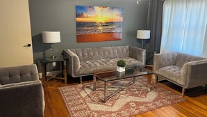 Una sala de estar con un sofá gris, dos sillones, una mesa de centro de vidrio y una alfombra estampada.