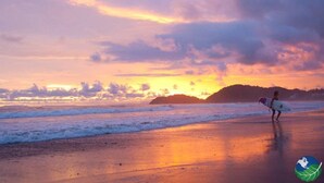A beach scene with a sunset, waves, and a person carrying a surfboard. - 11B Jaco Sol Quadplex - Luxury apartment near the beach and commerce (Jacó)