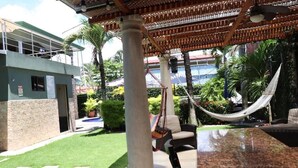A covered outdoor seating area with a hammock, a pool, and a building with a stone facade. - 11B Jaco Sol Quadplex - Luxury apartment near the beach and commerce (Jacó)