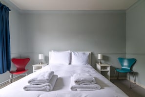 A hotel room with a bed, two nightstands, a red chair, a teal chair, and a window with curtains.