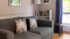 Een grijze bank met kussens met palmbladeren, een witte boekenkast met boeken, een vloerlamp en een raam met blauwe gordijnen.