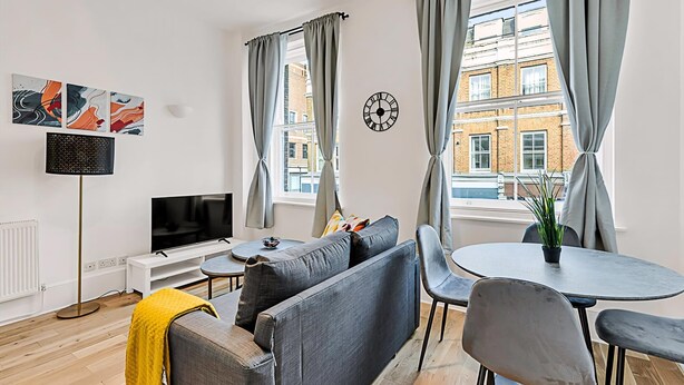 Een moderne woonkamer met een bank, televisie en eettafel, grote ramen met gordijnen en uitzicht op de stad.