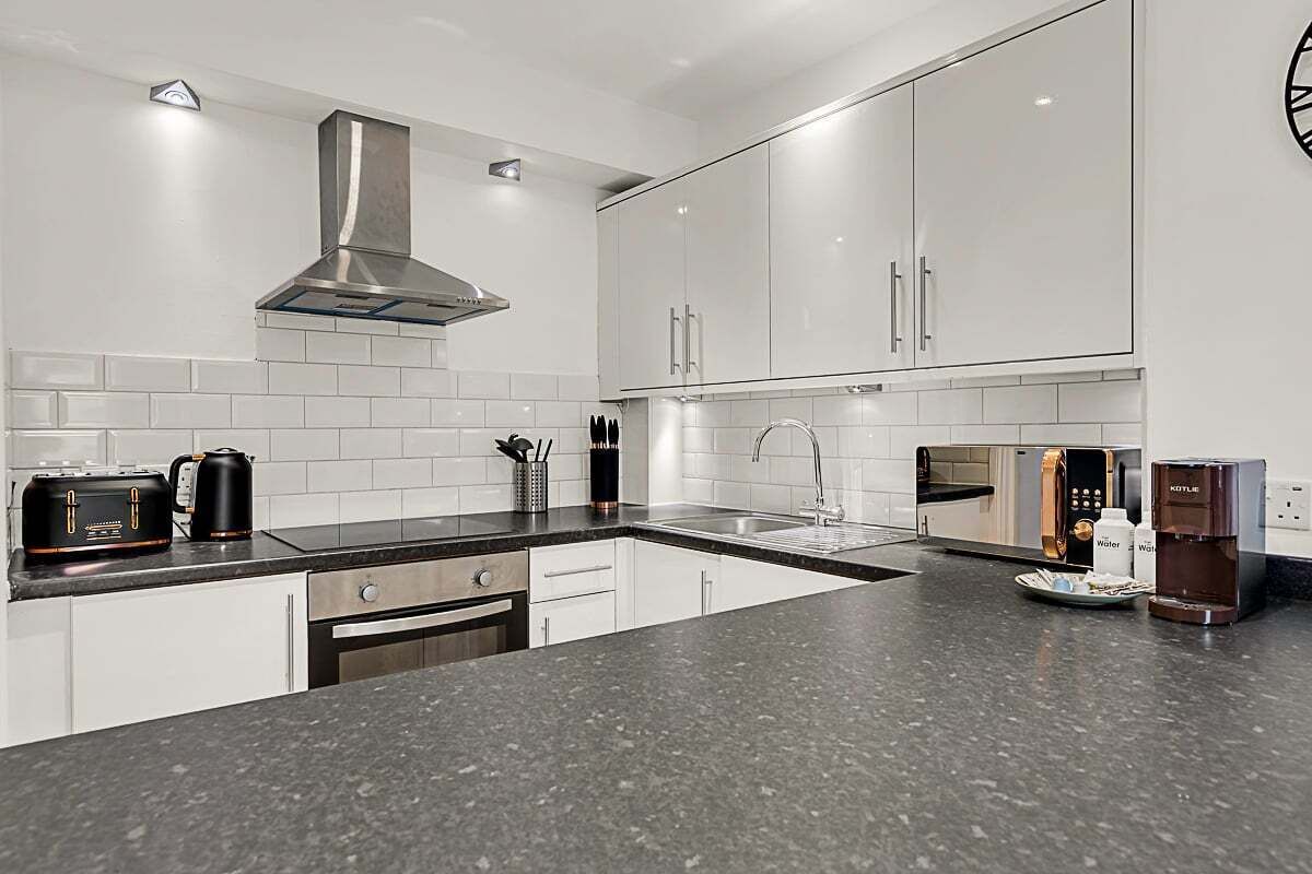 A modern kitchen with white cabinets, a stainless steel range hood, a built-in oven, a coffee machine, and a microwave.