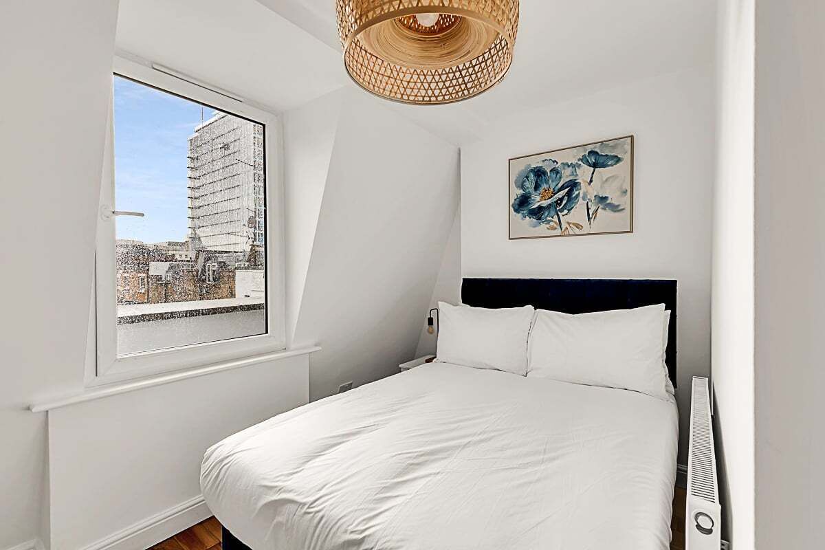 A bedroom with a bed, a window with a view of a building, a wall-mounted artwork, and a ceiling light fixture.