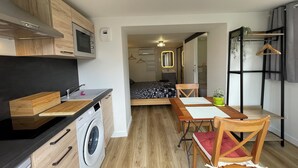 A compact apartment with a kitchenette, a bed with a patterned bedspread, a dining area with a table and chairs, and a small potted plant.