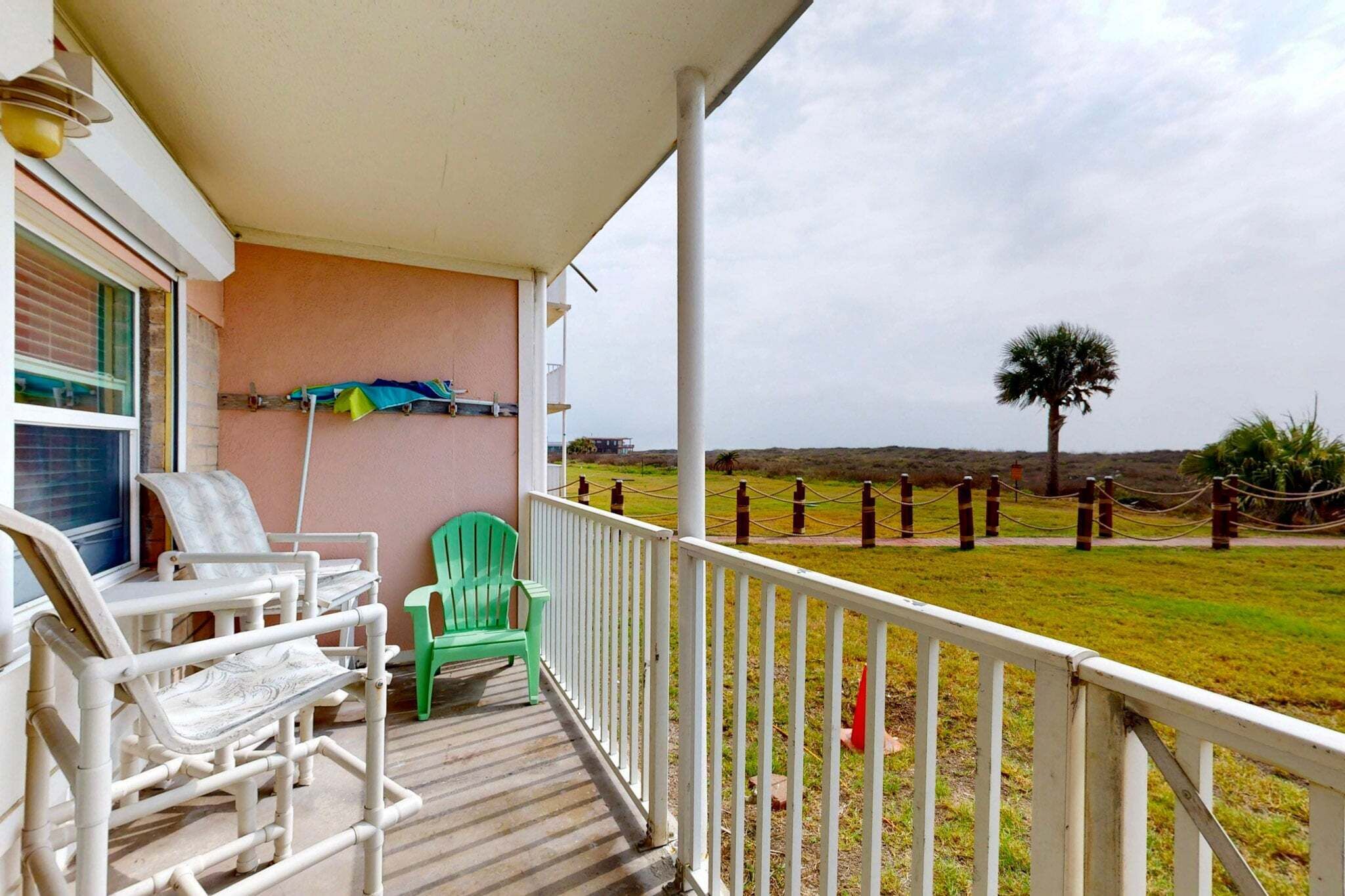 A balcony with chairs, a green chair, and a view of a fenced area with a tree.