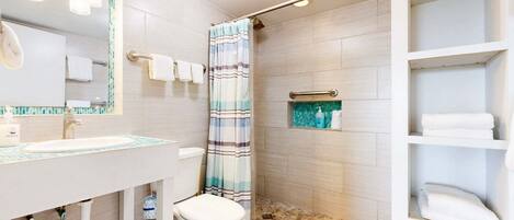 Une salle de bain équipée d’un lavabo, de toilettes, d’une douche et d’étagères où sont rangées les serviettes.