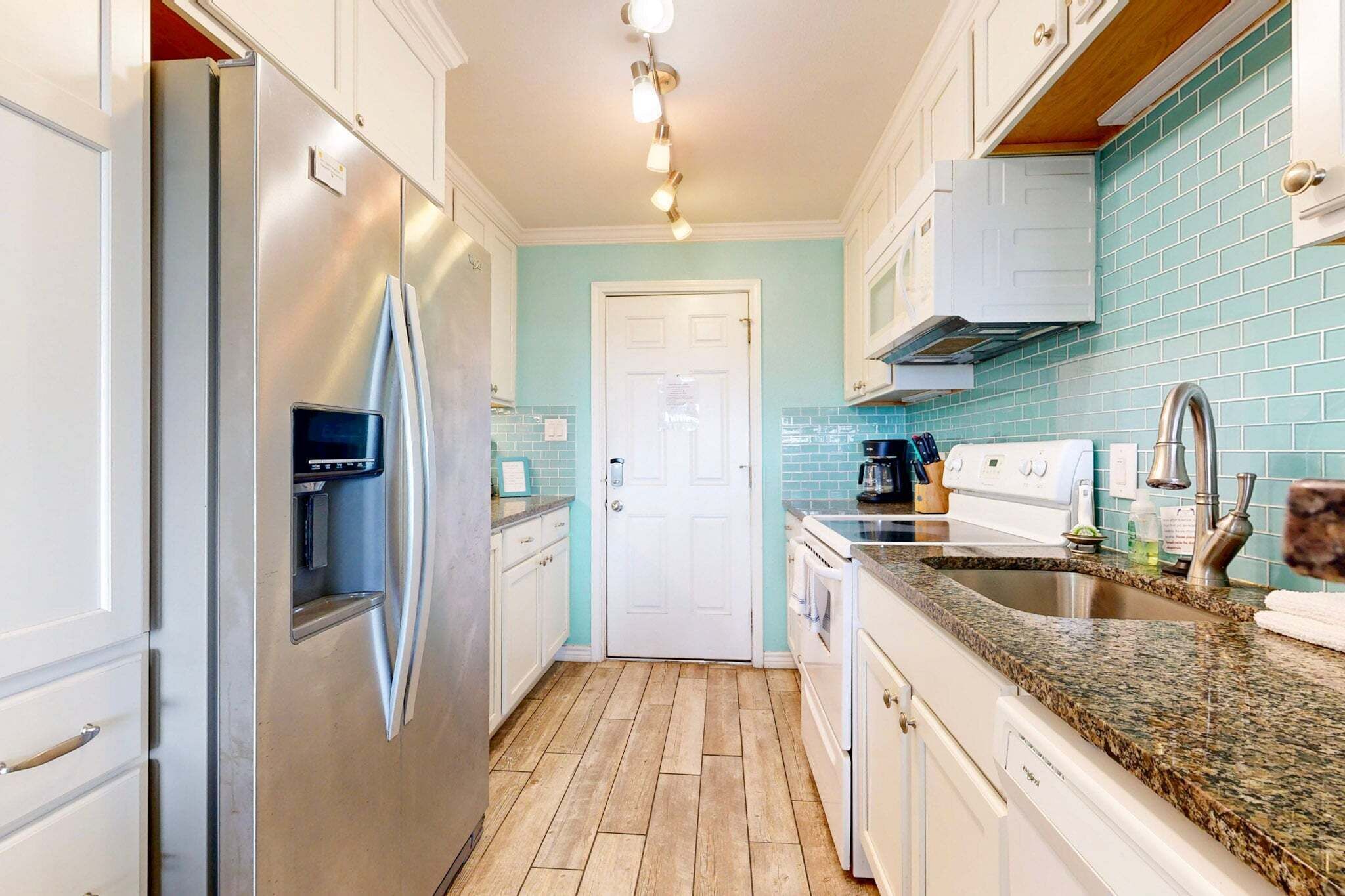 A kitchen with a stainless steel refrigerator, white cabinets, a teal backsplash, and a granite countertop.