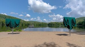 Una playa con dos sombrillas verdes, una orilla arenosa, un lago tranquilo y una colina boscosa al fondo.