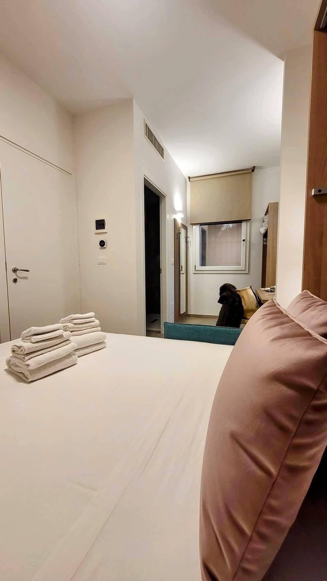 A hotel room with a bed, a chair, a stack of towels, a door, and a window.
