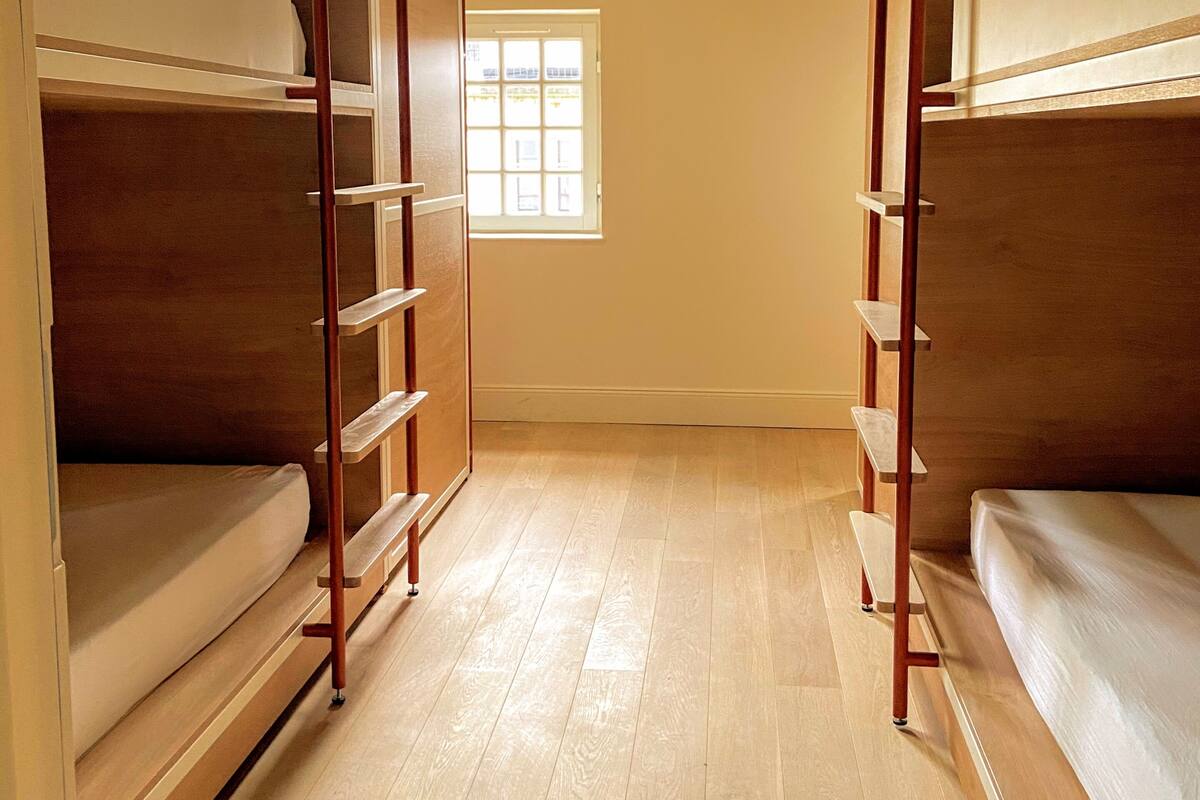 A dormitory room with bunk beds, wooden flooring, a skylight, and a window.