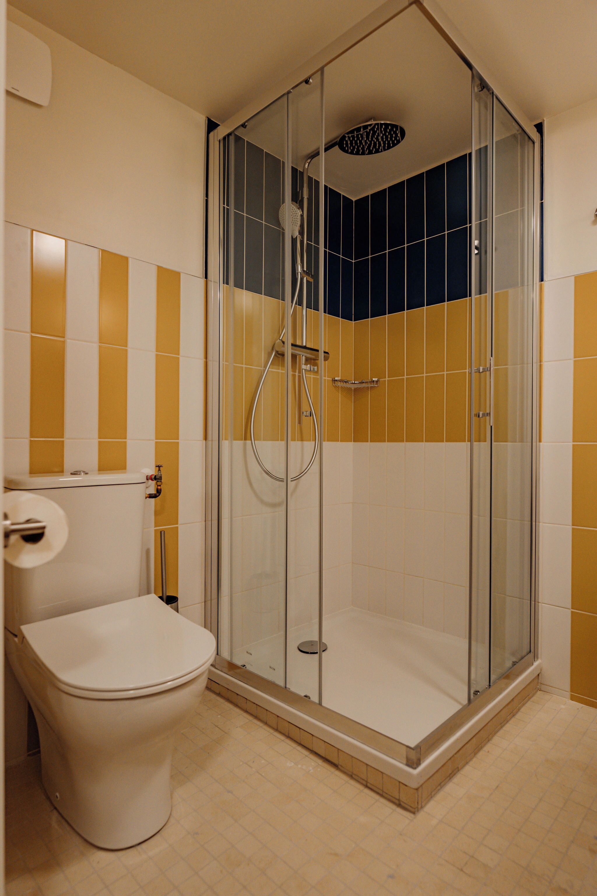 Een badkamer met een douche met glazen wand, een toilet en gele en witte tegels op de muren.