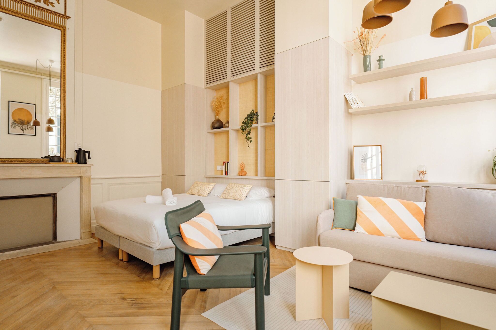 A modern hotel room with a bed, a sofa, a chair, a small table, and a shelf with decorative items.