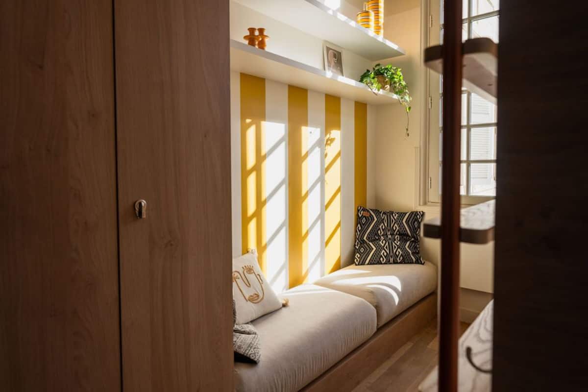 A room with a wooden wardrobe, a sofa with patterned cushions, and a shelf with decorative items.