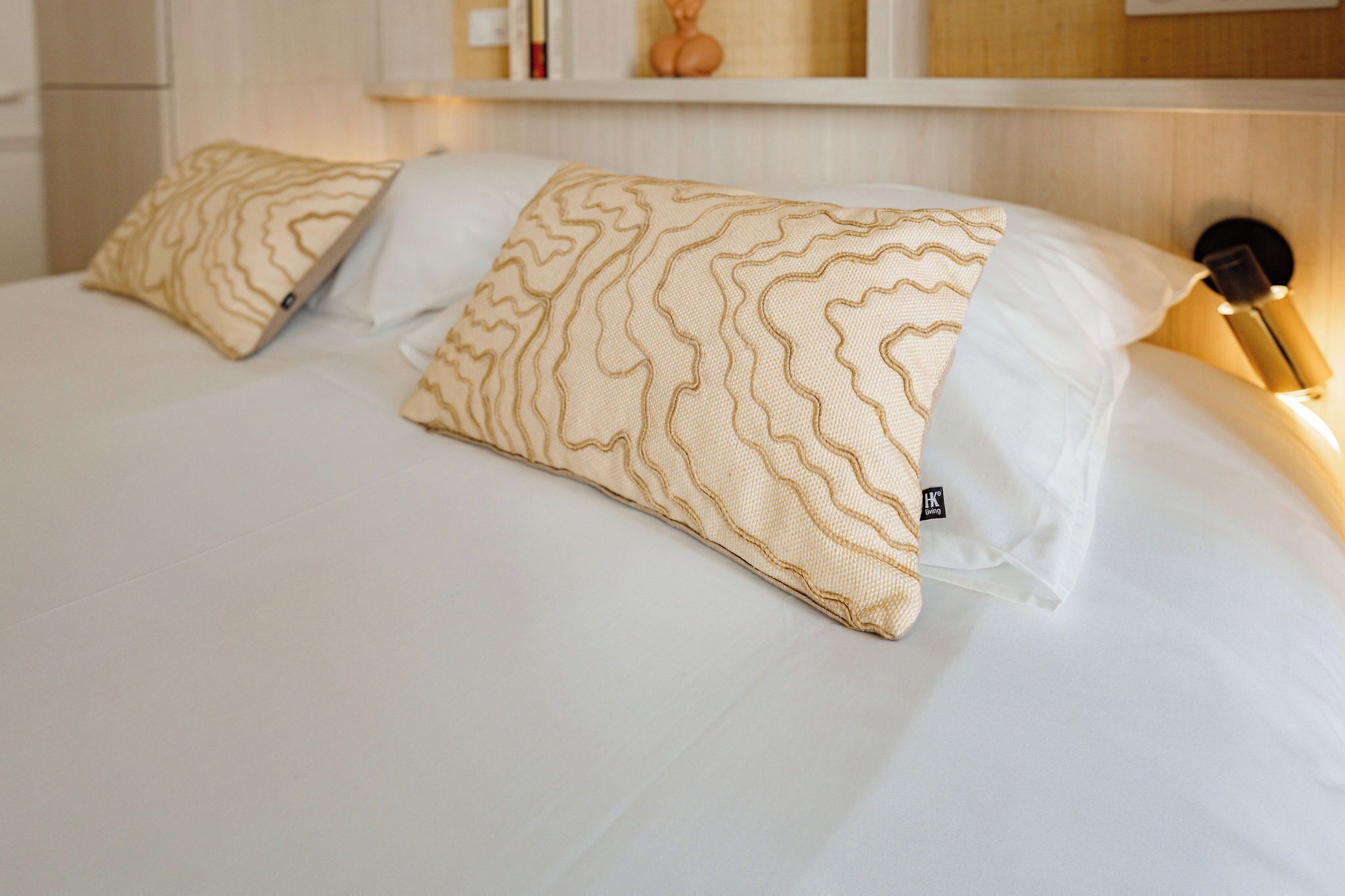 A neatly made bed with two patterned pillows and a white pillow, a bedside lamp, and a shelf with books and a decorative item in the background.