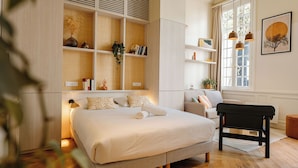 A bedroom with a large bed, a bedside table, a chair, a bookshelf, and a window with curtains. - CENTRAL (La Rochelle)