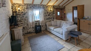 A cozy room with a stone wall, a wooden ceiling, a sofa, a small table, and a kitchen area. - Elegant and Secluded 1-bed Cottage Near Bideford (Bideford)