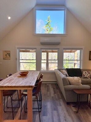 A spacious room with a high ceiling, a dining table with chairs, a sofa, and large windows offering a view of trees and a clear sky.