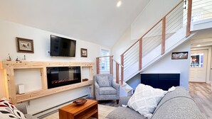 A spacious living room with a large grey sofa, a wooden coffee table, and a fireplace. A staircase leads to an upper level, and a television is mounted on the wall. - Brand New, Private Home w/ Nordic Hot Tub- Just 1/2 mile to Jay Peak Resort (Jay)