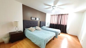 Une chambre avec deux lits, un ventilateur de plafond, une table de chevet avec une lampe et une fenêtre avec des rideaux.