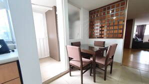 Un coin repas avec une table en verre et quatre chaises, un séparateur en bois avec un motif quadrillé, et une vue sur un espace de vie comprenant un canapé et une lampe.