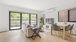A modern living room with a sofa, a dining table with chairs, a TV, and a large window with a view of a wooden fence and greenery. - StayAU Modern 4BRM TH Bayswater (Bayswater North)