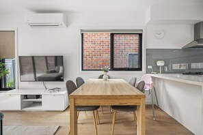 A modern kitchen with a brick wall, a dining area with a wooden table and chairs, and a TV on a white cabinet. - StayAU Modern 4BRM TH Bayswater (Bayswater North)