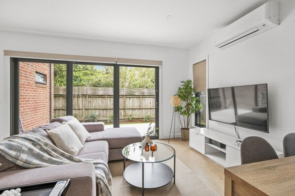 A modern living room with a large TV, a sofa, a coffee table, and a view of a backyard. - StayAU Modern 4BRM TH Bayswater (Bayswater North)