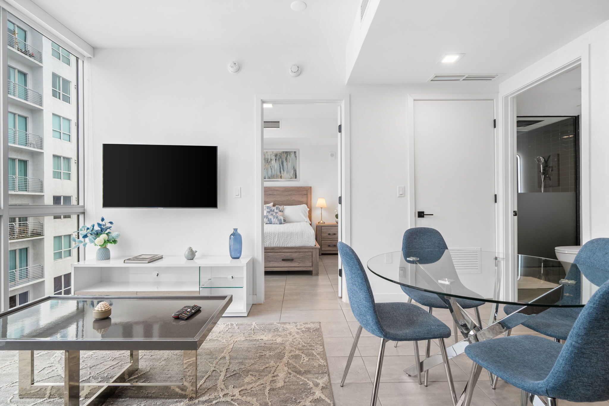 Een moderne hotelkamer met een flatscreen televisie, een glazen eettafel met stoelen, een bed met een houten hoofdbord en een badkamer met douche.