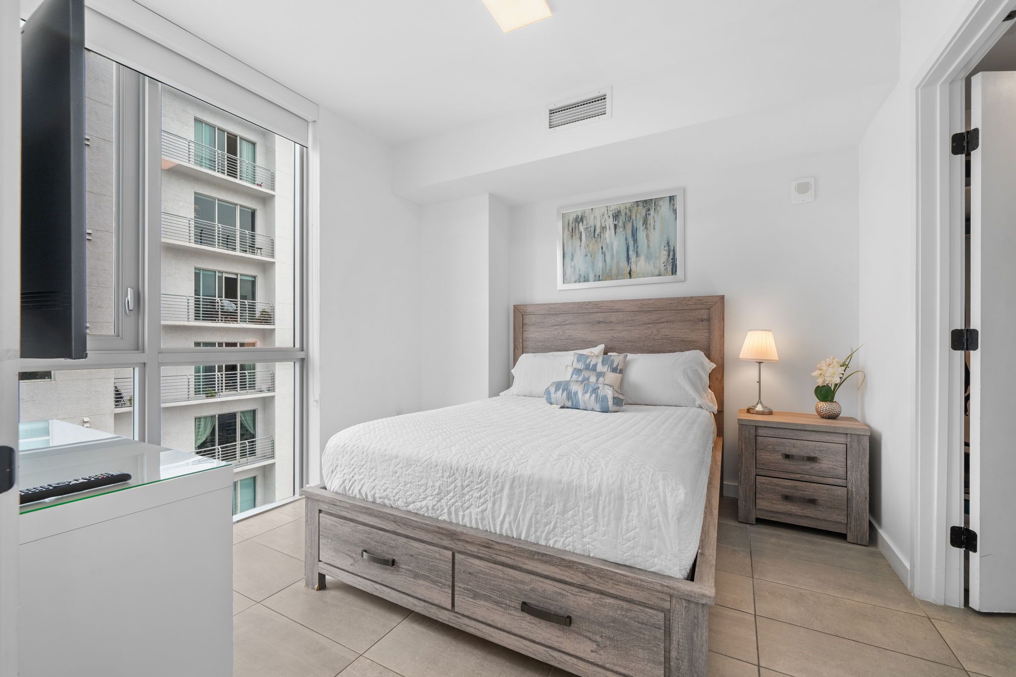 Un dormitorio con cama, mesita de noche, lámpara y vista al balcón exterior.