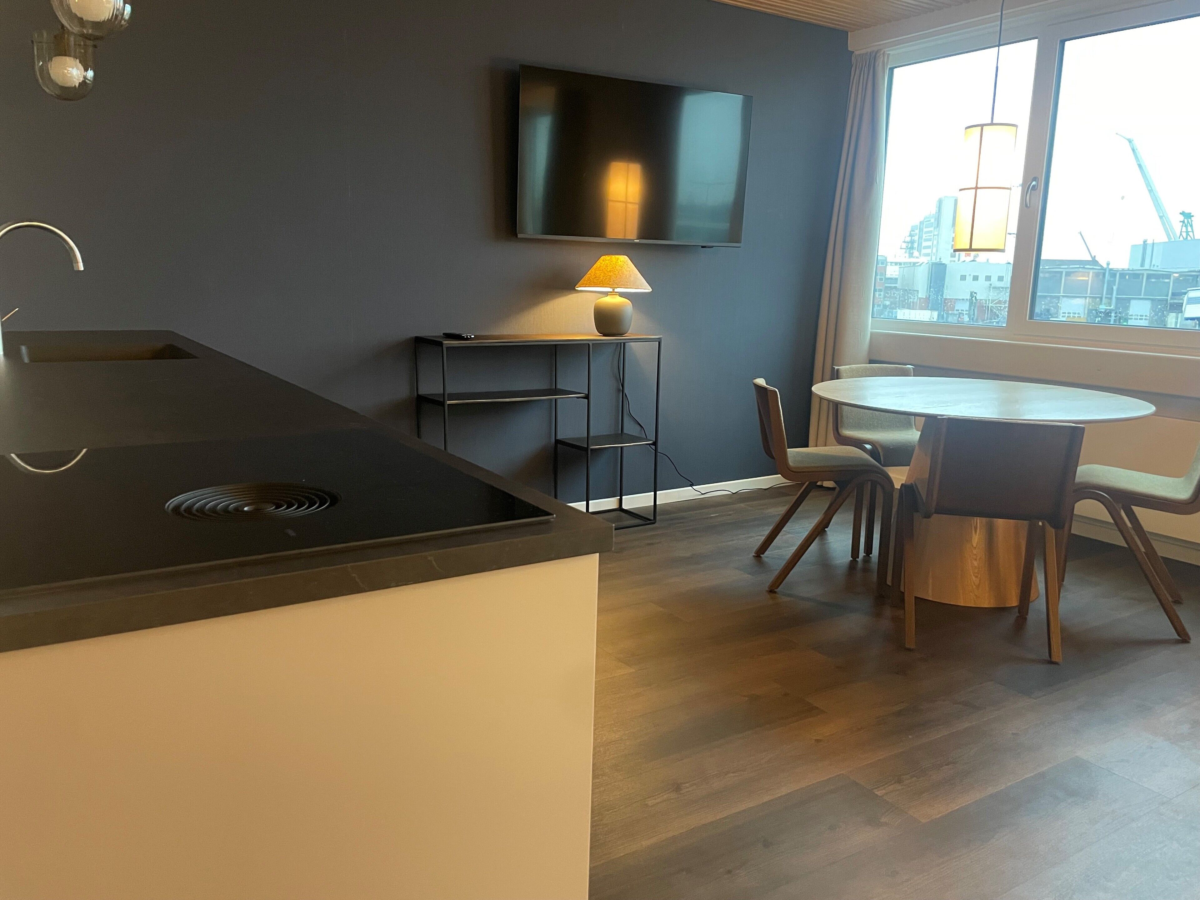 Une cuisine moderne dotée d’une cuisinière intégrée, un coin repas avec une table ronde et des chaises, un téléviseur fixé au mur et une fenêtre offrant une vue sur le paysage urbain.