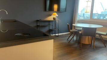 Une cuisine moderne dotée d’une cuisinière intégrée, un coin repas avec une table ronde et des chaises, un téléviseur fixé au mur et une fenêtre offrant une vue sur le paysage urbain.